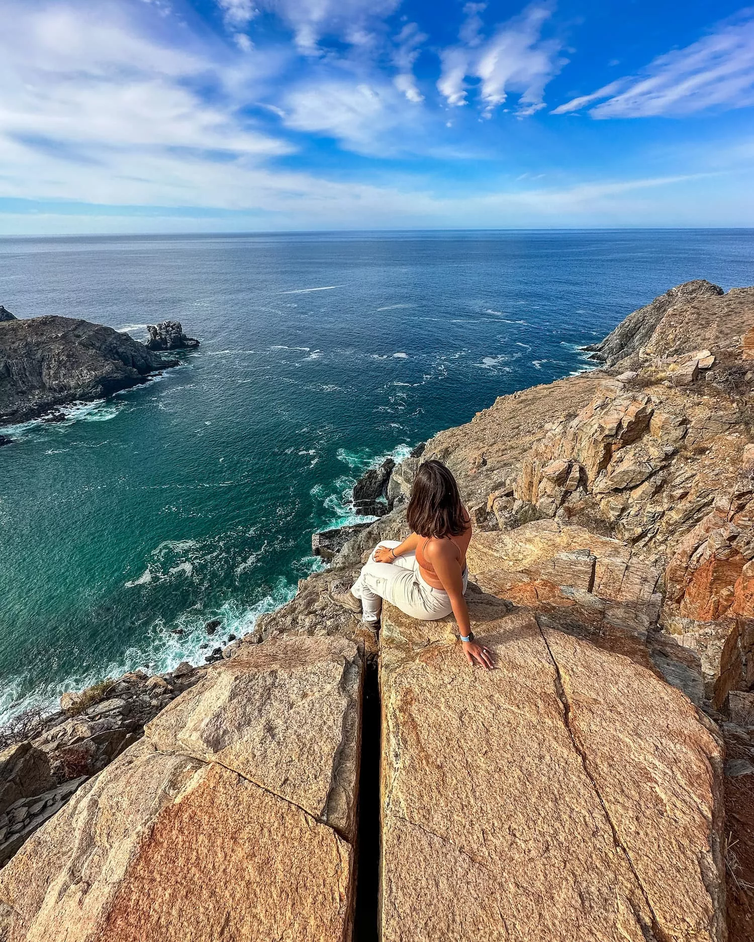 girl sitting on the edge of a cliff with water beyond her punta lobos trail peak