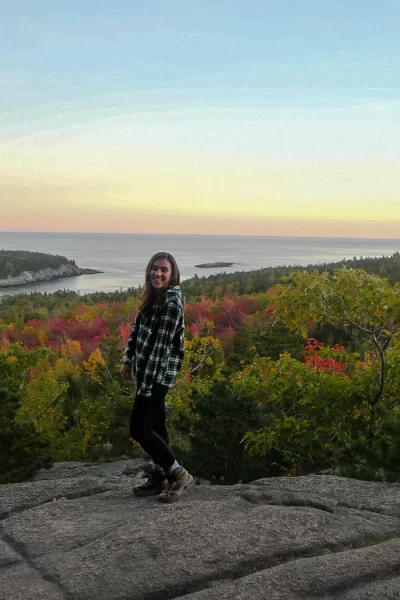 girl standing and smiling on beehive trail lookout in acadia national park with fall colored trees behind