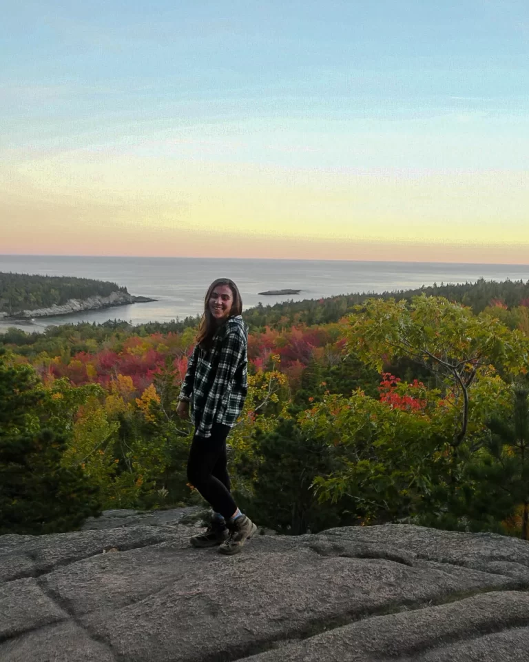 girl standing and smiling on beehive trail lookout in acadia national park with fall colored trees behind