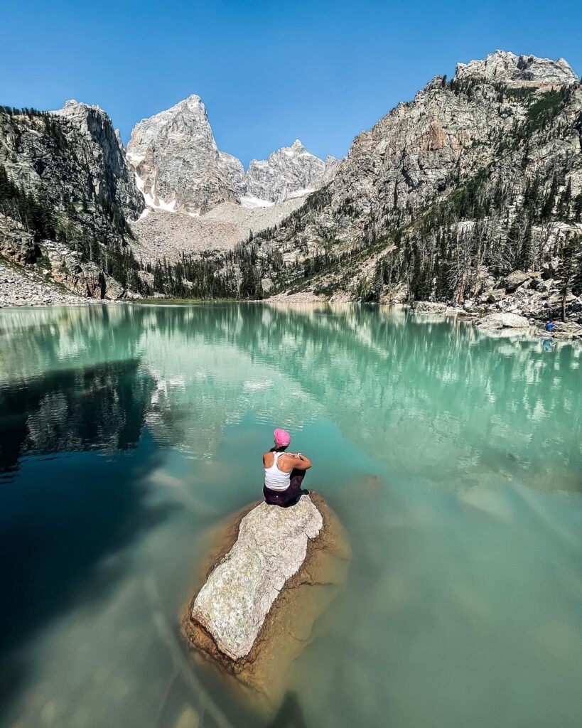 girl sitting on a rock with turquoise lake all around and teton mountains behind her
