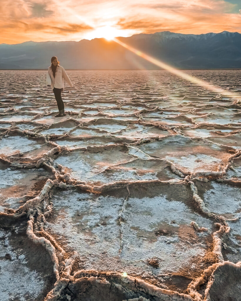 girl stands on salt flats of badwater basin as sun sets behind her with an orange glow