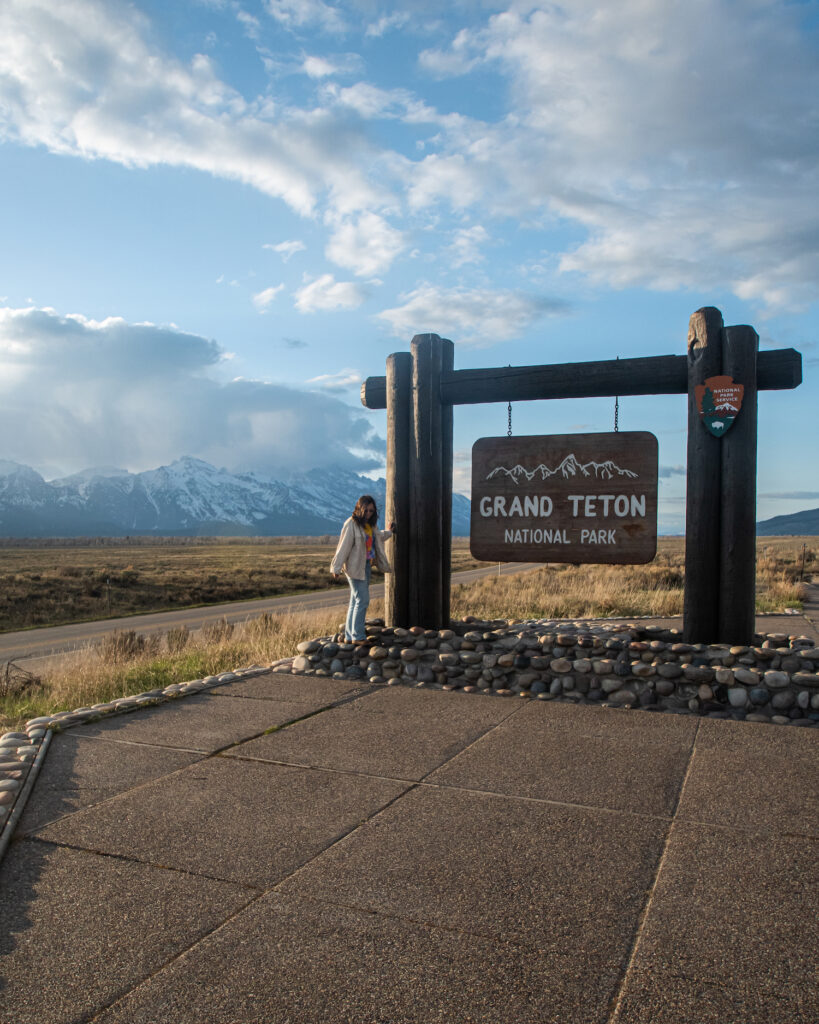 Grand teton sign in front of the mountains with girl standing next to the sign