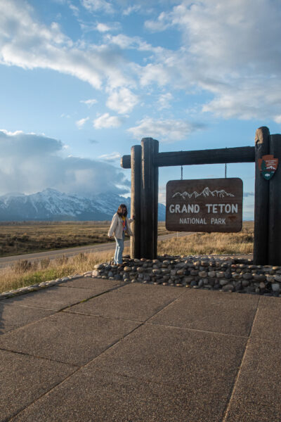 girl standing next to grand teton park sign with. mountains in back