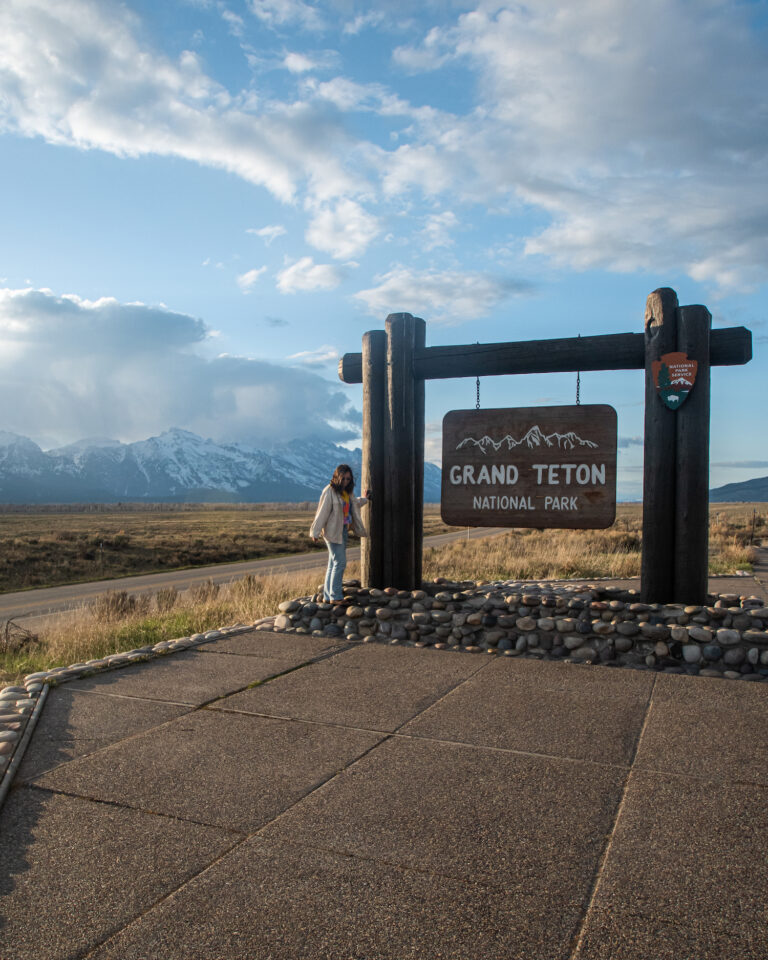 girl standing next to grand teton park sign with. mountains in back