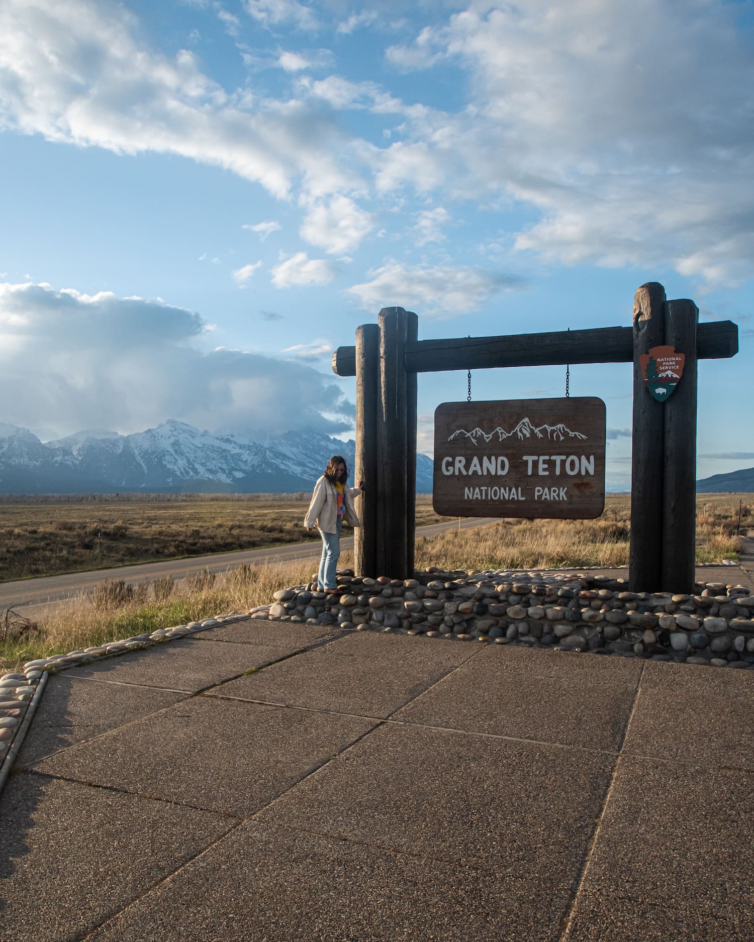 girl standing next to grand teton park sign with. mountains in back