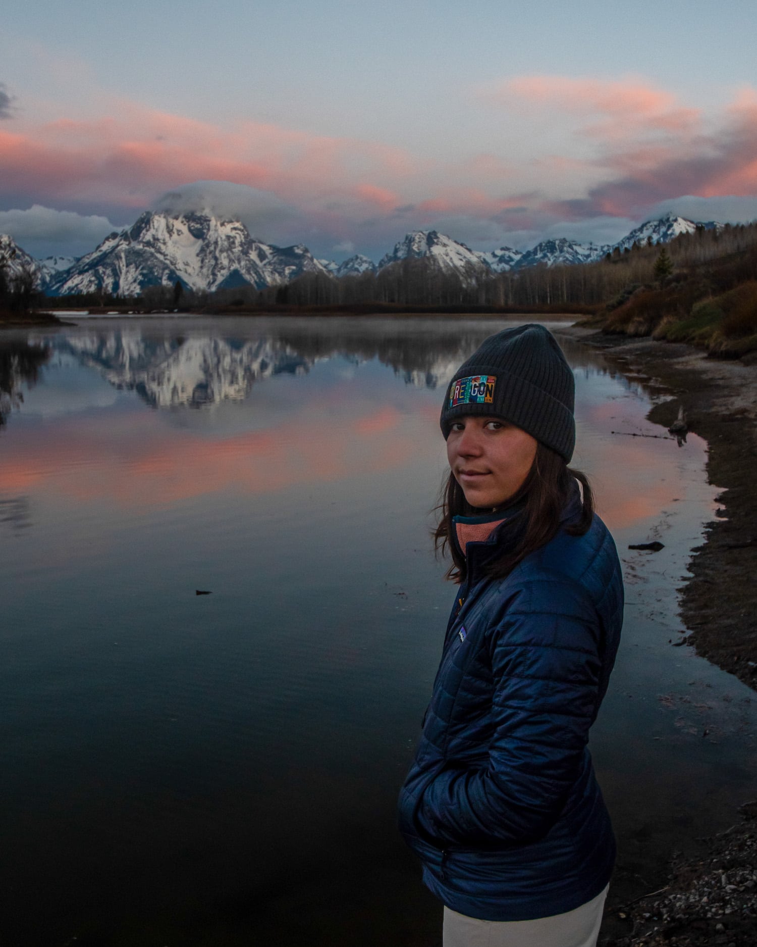 girl standing in front of the Grand Tetons as sun rises and the mountains reflect with pink clouds at Oxbow Bend