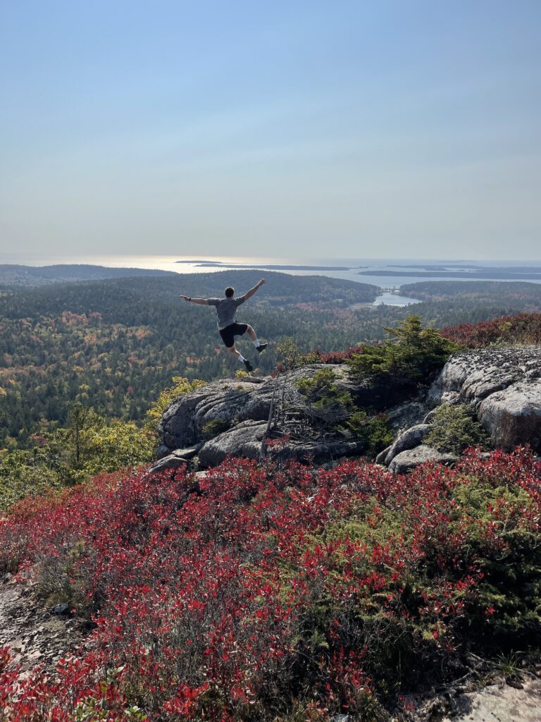 boy jumping up and kicking his feet on a mountain with red bush in front of him
