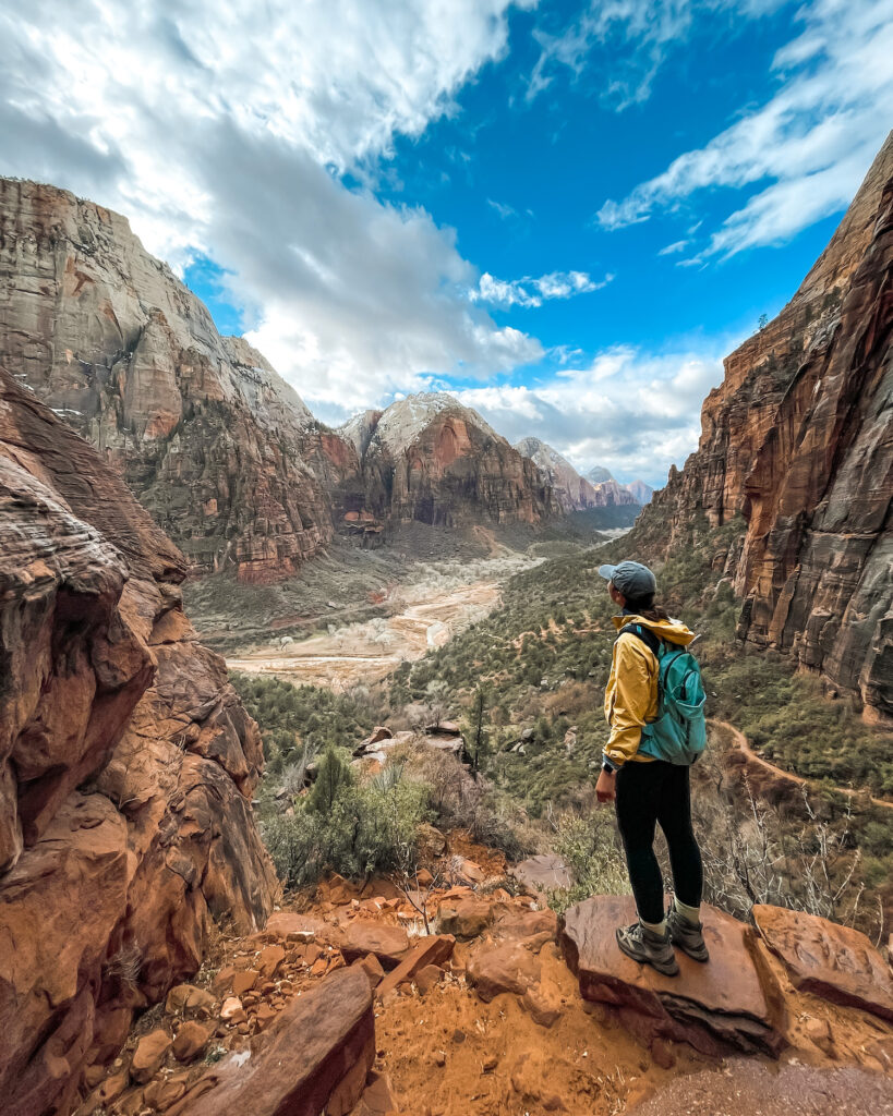 girl standing on red cliff of Angels Landing with mountains and river out in front of her (her back is to the camera)
