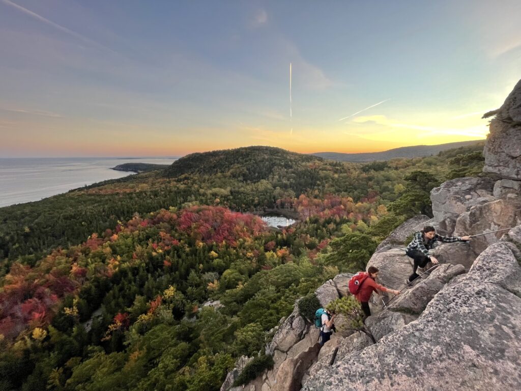 people climbing up rungs on a mountain with sun setting and reds and oranges in the trees