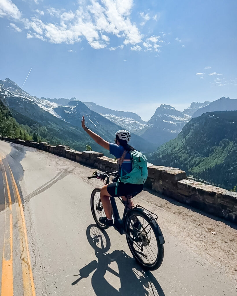 girl biking one handed with arm up as she goes up going to the sun road with mountains behind her