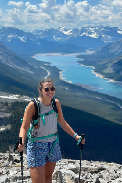 girl smiling out with poles in her hands as she stands on a mountain summit with lake behind her and mountains
