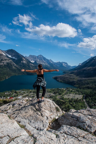 girl standing on a mountain with arms out while blue lakes of Waterton are below her and mountains around her