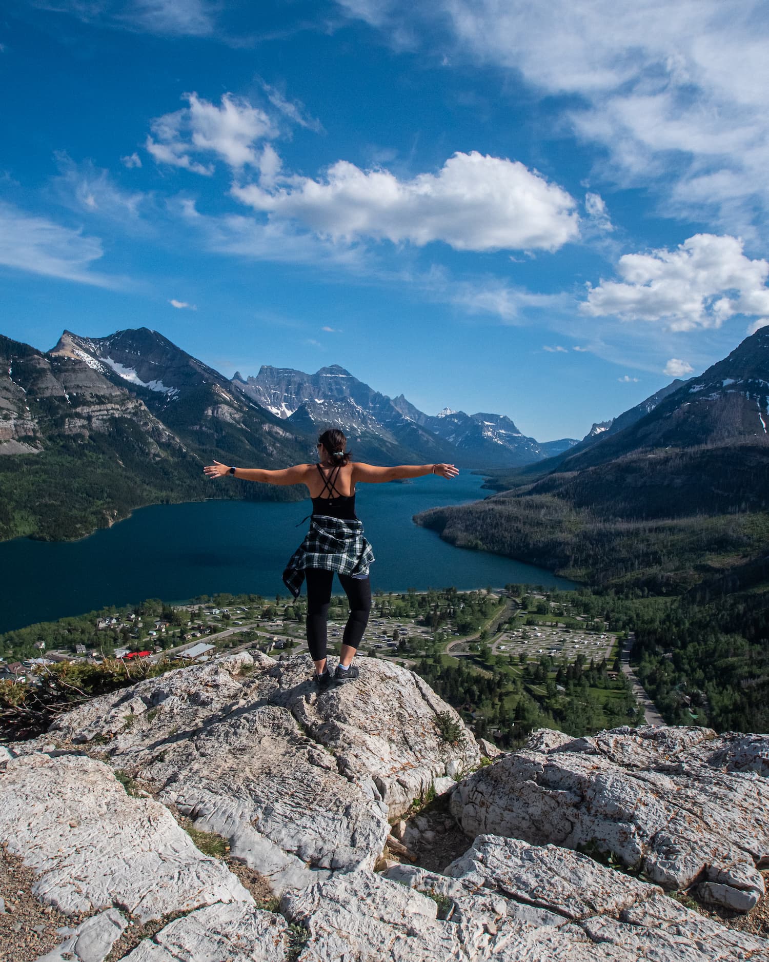 girl standing on a mountain with arms out while blue lakes of Waterton are below her and mountains around her