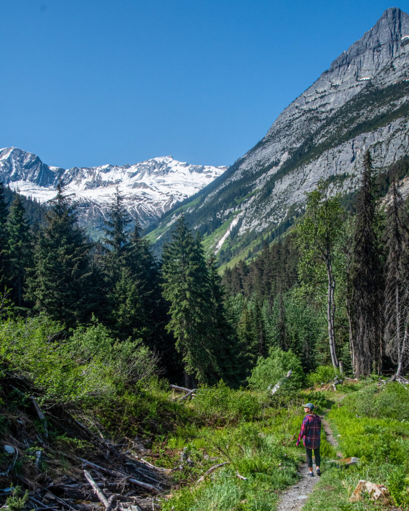 girl hiking on a trail with tall mountain peaks behind her and a forest