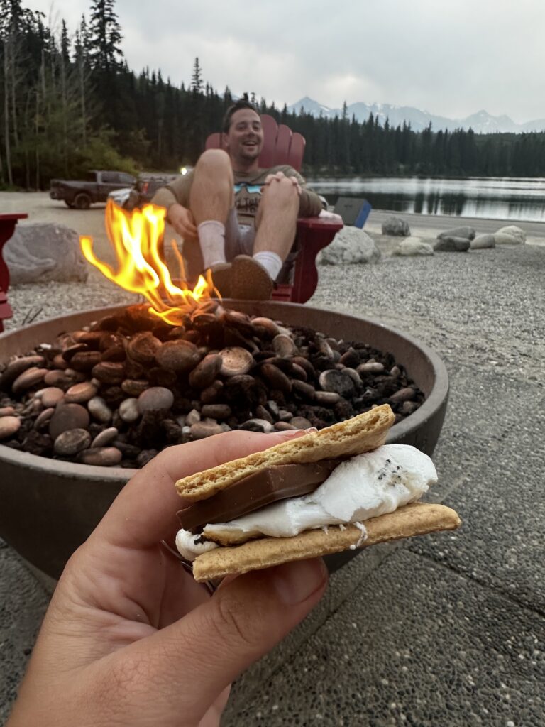 Smores in front of a fire with boy sitting on a chair in front of lake. 