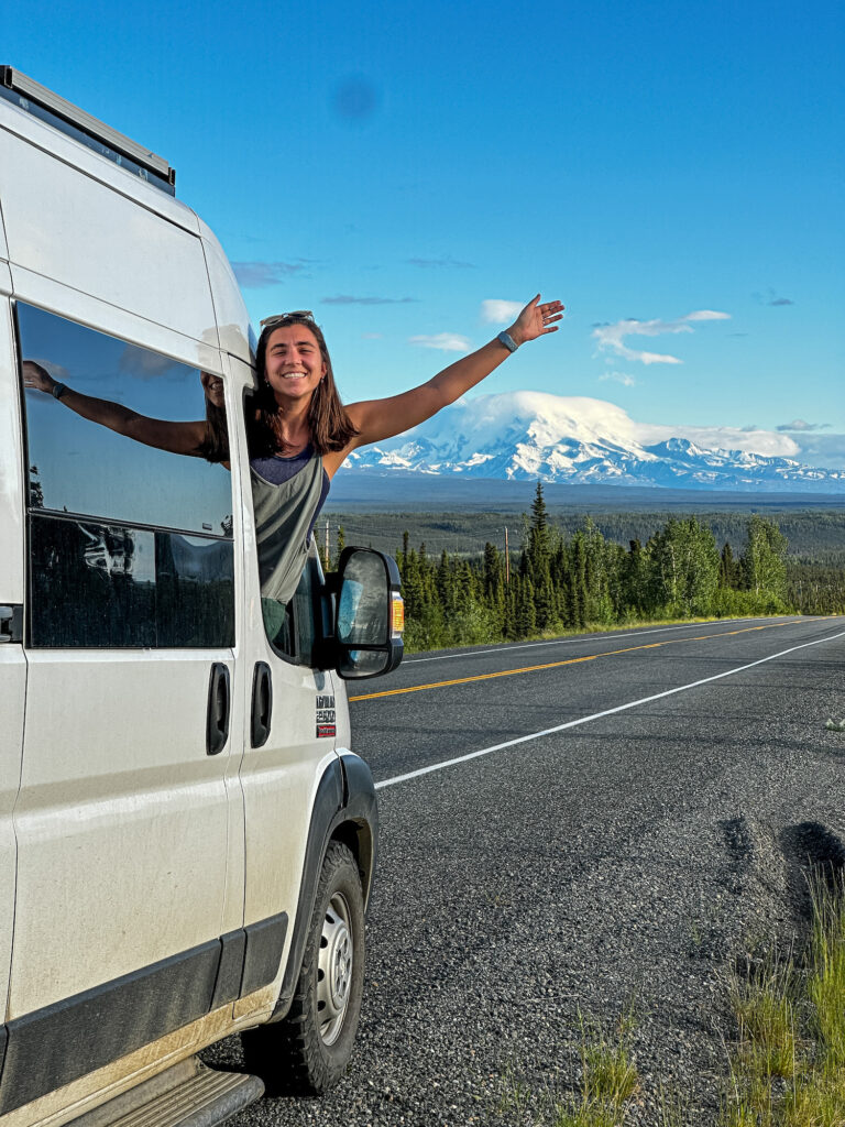 girl smiling with her hand out the window of her van and Alaska Mountains behind her