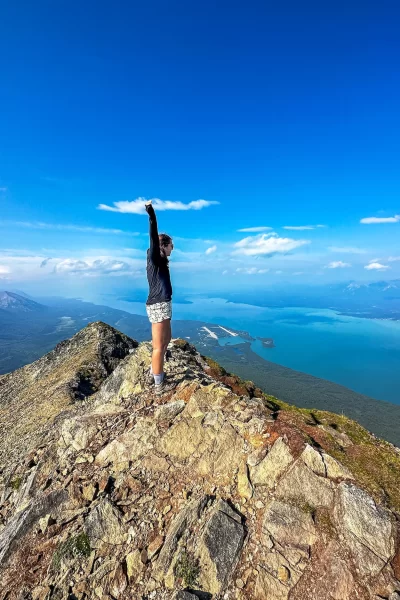 girl with arms up on the top of Tanalian Moutnain peak with bright blue watrs of Lake Clark below her