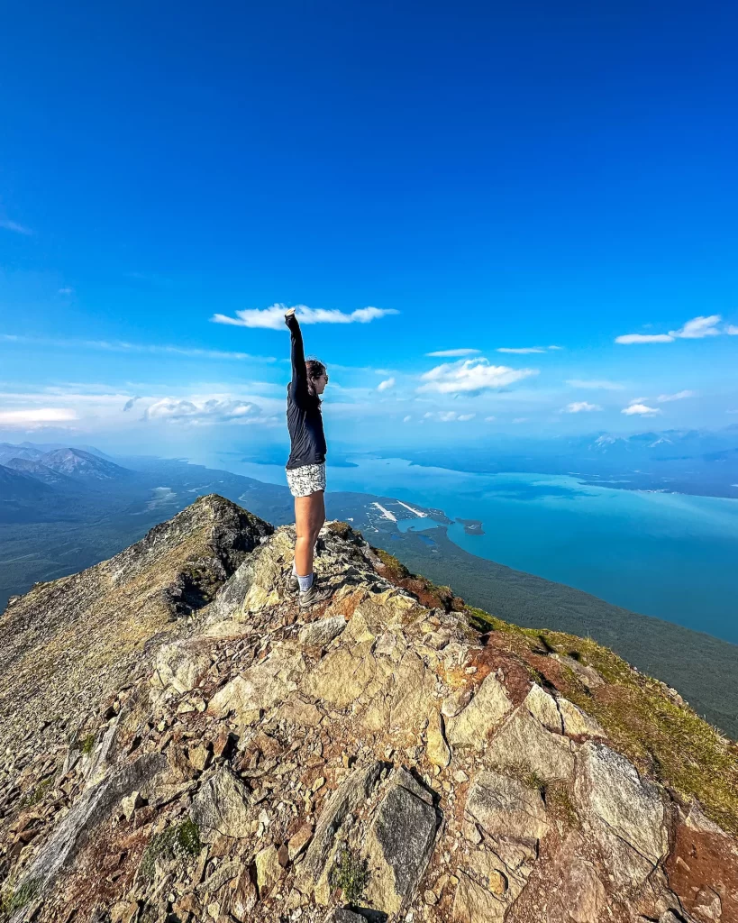 girl with arms up on the top of Tanalian Moutnain peak with bright blue watrs of Lake Clark below her