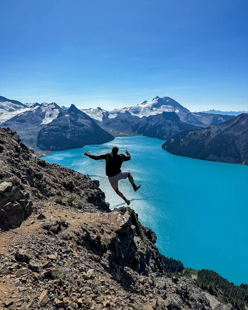 boy jumping on the top of a mountain summit with bright blue lake behind him