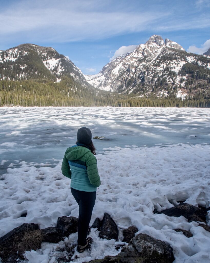girl in a jacket on the shore of ice covered lake with tetons behind it