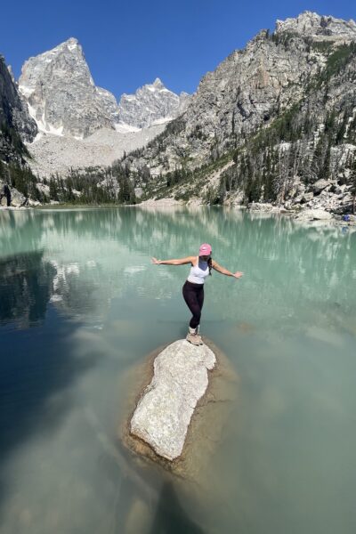 woman standing on a rock in turquoise lake with grand tetons behind