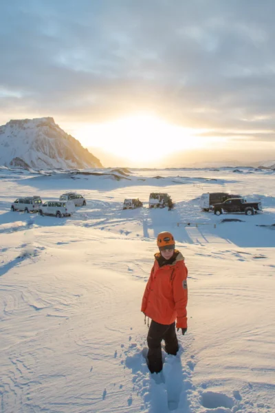 Woman standing on the ice in iceland with a mountain behind her and super jeeps
