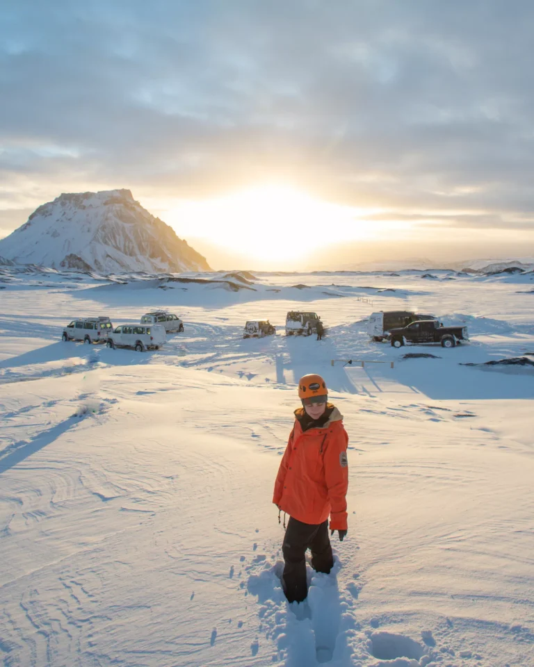 Woman standing on the ice in iceland with a mountain behind her and super jeeps