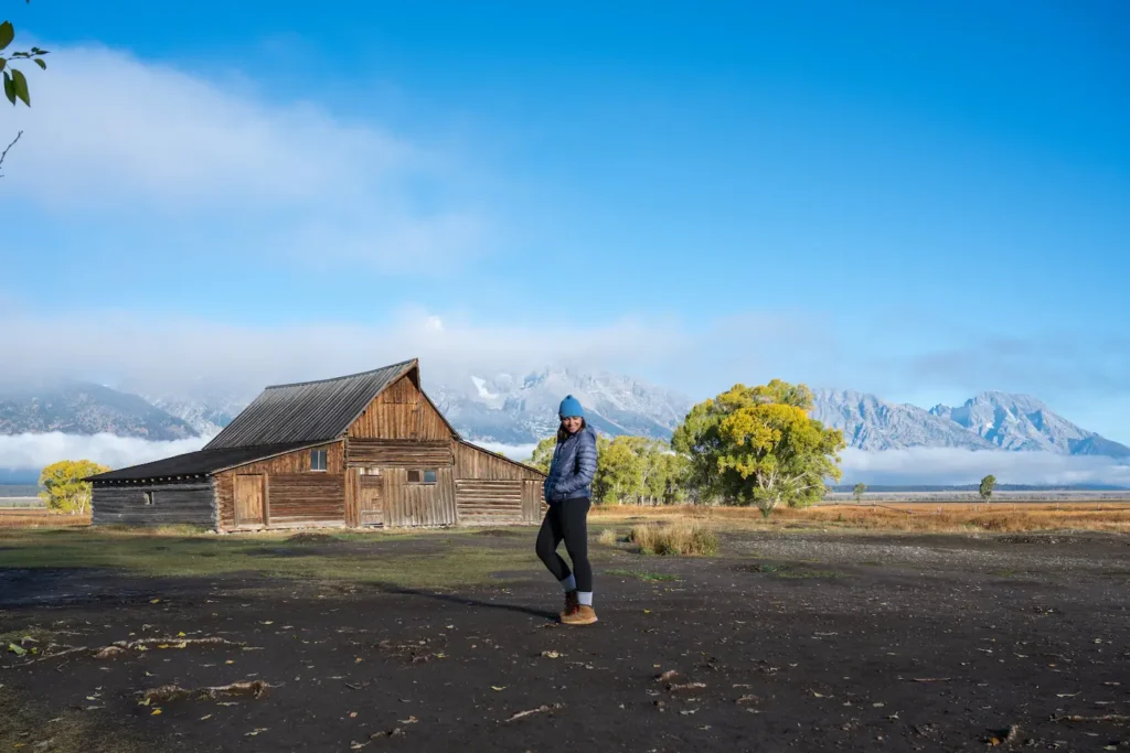 girl standing in front of Mormon Row Cabin and tetons wtih hands in pockets of her jacket