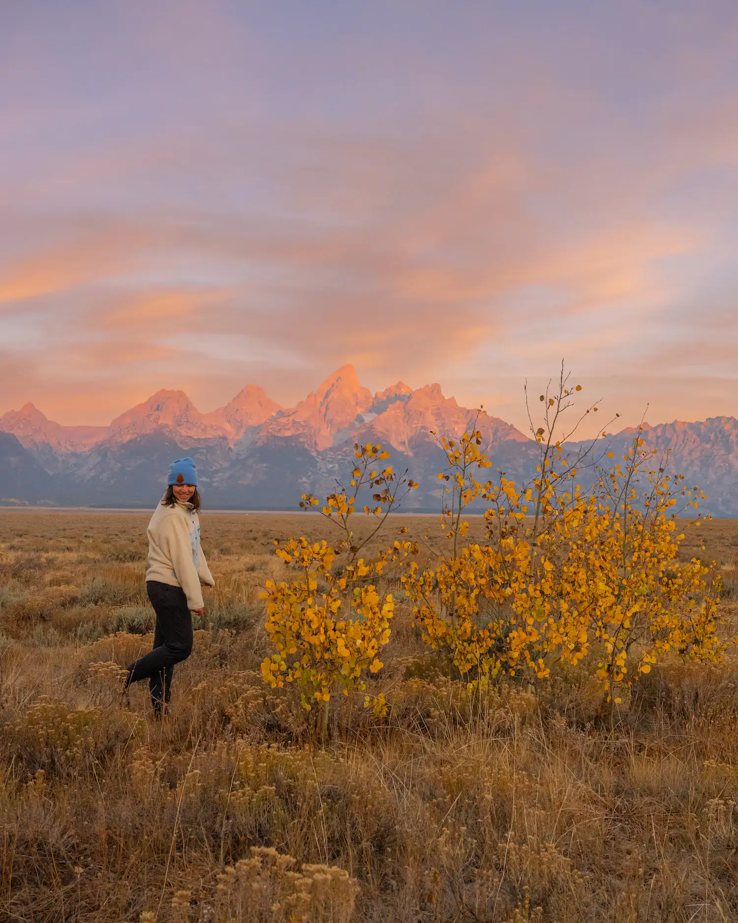 Girl standing in front of tetons at sunrise with fall colors on a tree next to her