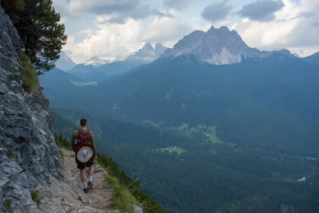 Guy walking on the mountain side with railing on left and sharp drop off to right and mountains in background